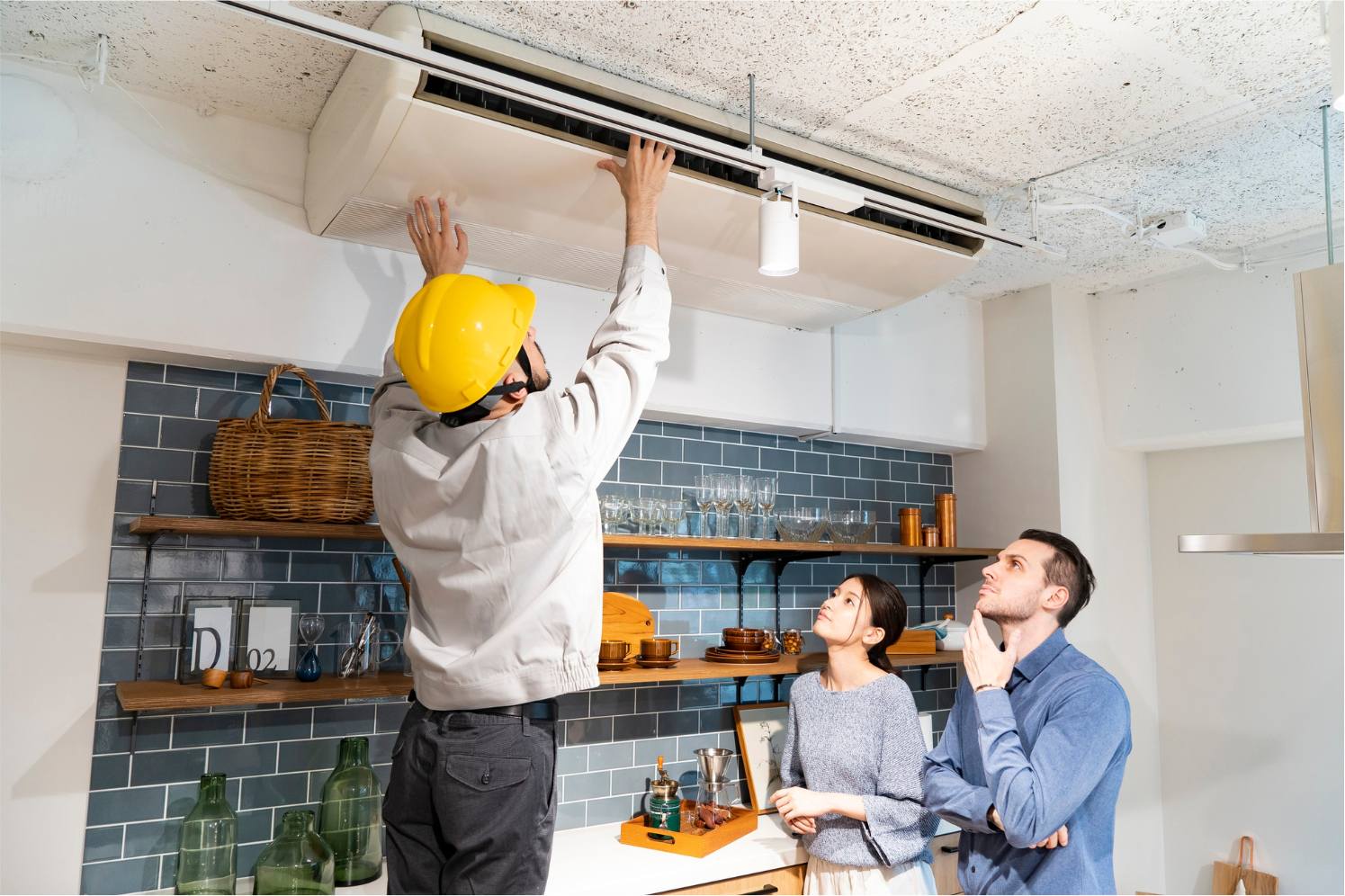 Technician installing a ceiling light fixture while two people watch in a kitchen