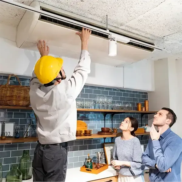 Technician installing a ceiling light fixture while two people watch in a kitchen