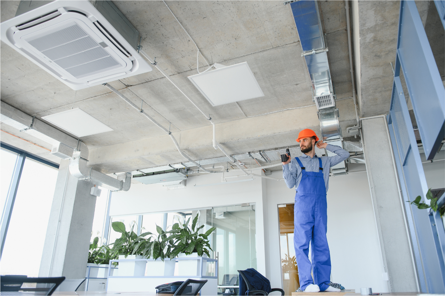 Worker in blue overalls and orange hard hat repairing ceiling in a bright office.