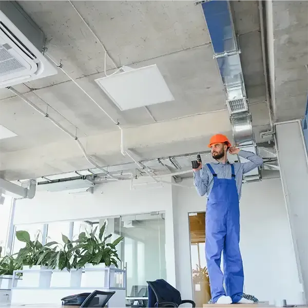 Worker in blue overalls and orange hard hat repairing ceiling in a bright office.
