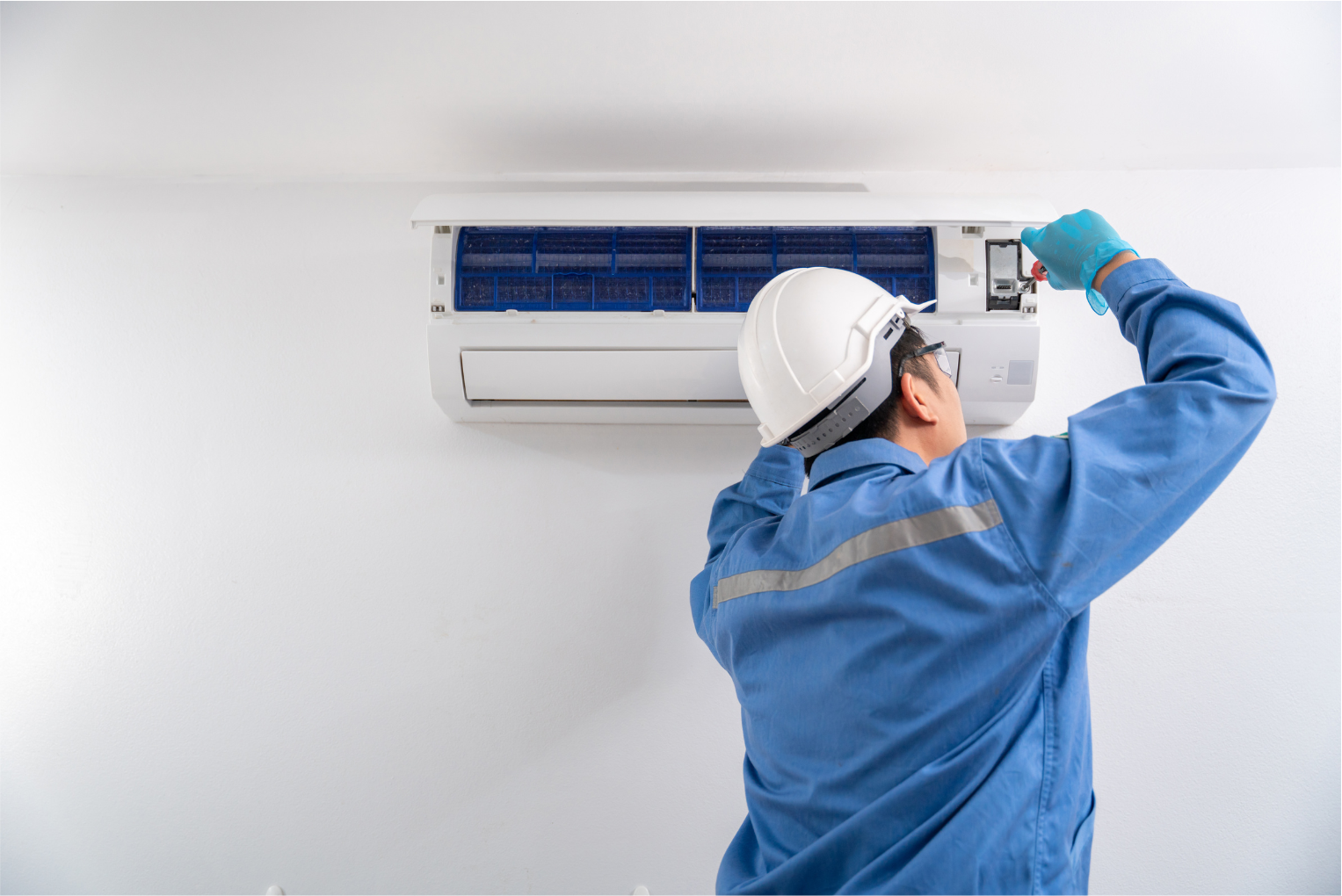 Technician servicing a wall-mounted air conditioner with tools, wearing a blue uniform and hard hat