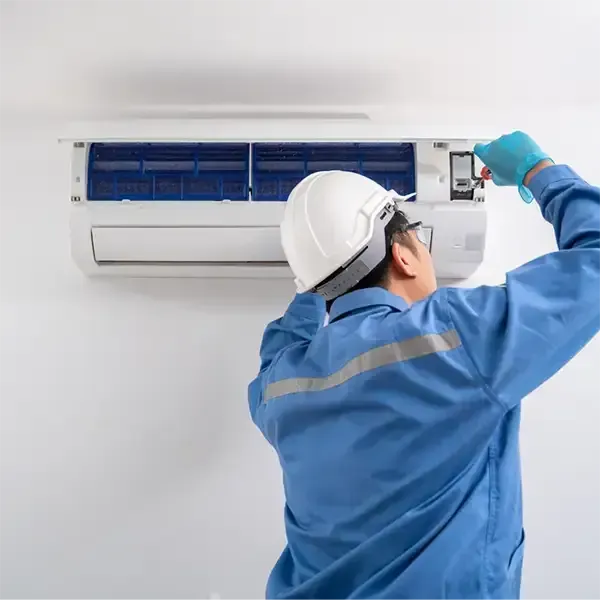 Technician servicing a wall-mounted air conditioner with tools, wearing a blue uniform and hard hat
