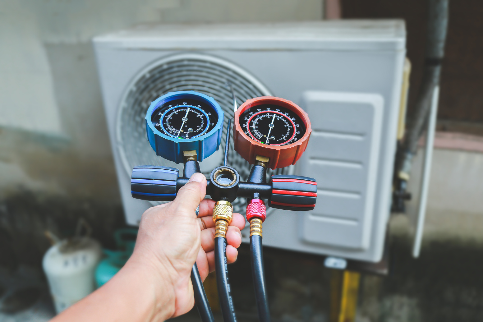 Hand holding blue and red HVAC manifold gauges in front of an outdoor AC unit