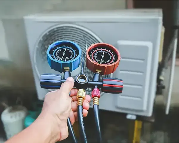 Hand holding blue and red HVAC manifold gauges in front of an outdoor AC unit