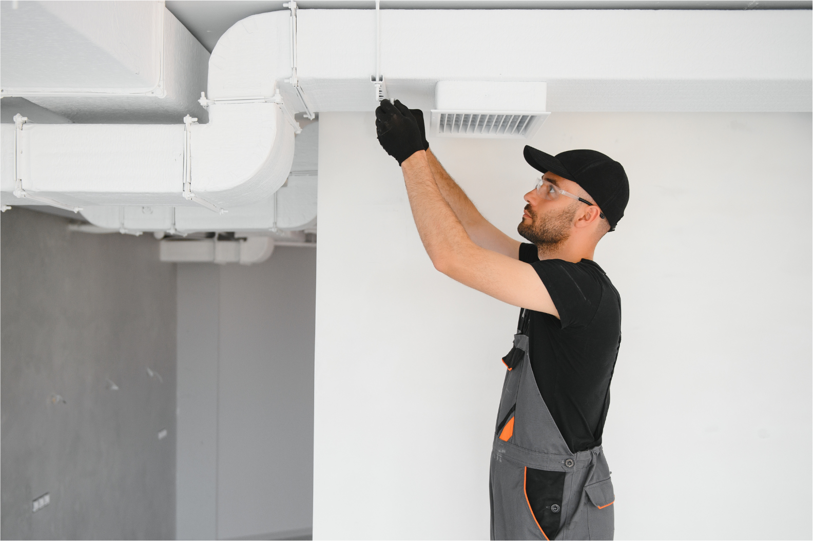 Technician inspecting white ceiling ductwork in a bright room, wearing black cap and work clothes