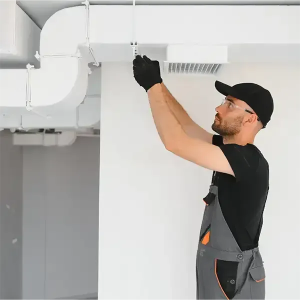 Technician inspecting white ceiling ductwork in a bright room, wearing black cap and work clothes