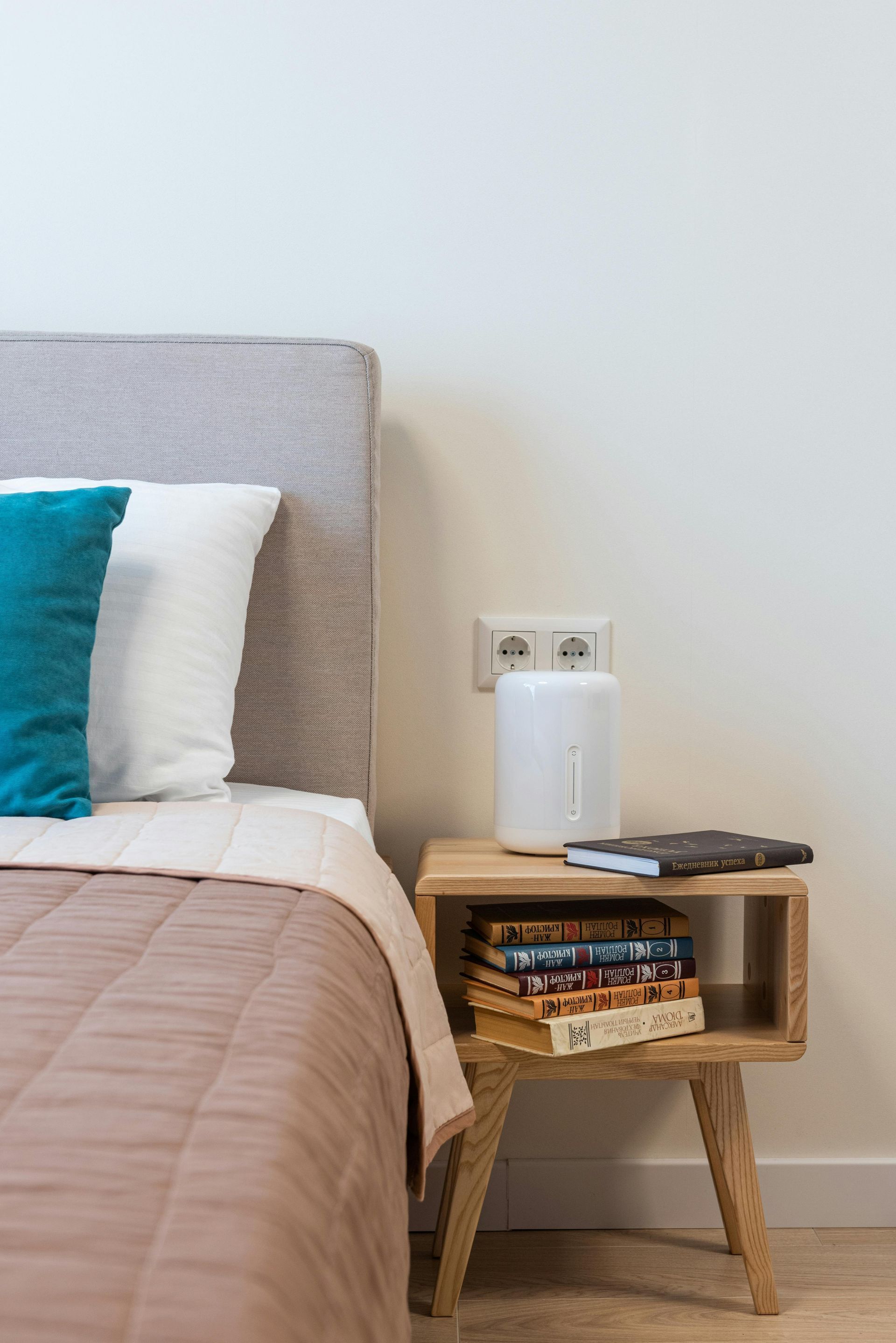 Bedroom corner with bed, bedside table, books, and a white appliance against a plain wall