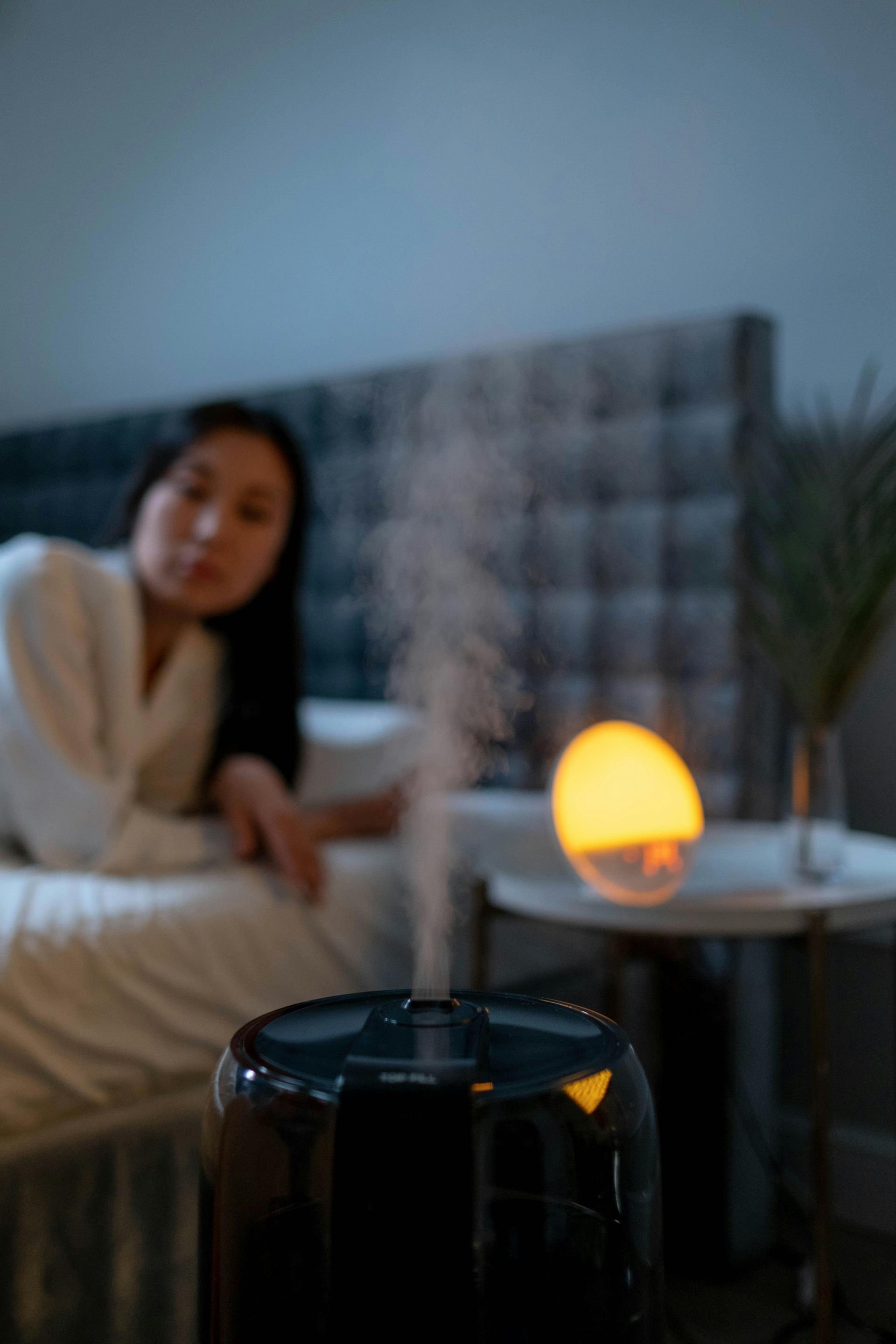 Woman lying in bed beside a glowing lamp, with a dark humidifier in the foreground