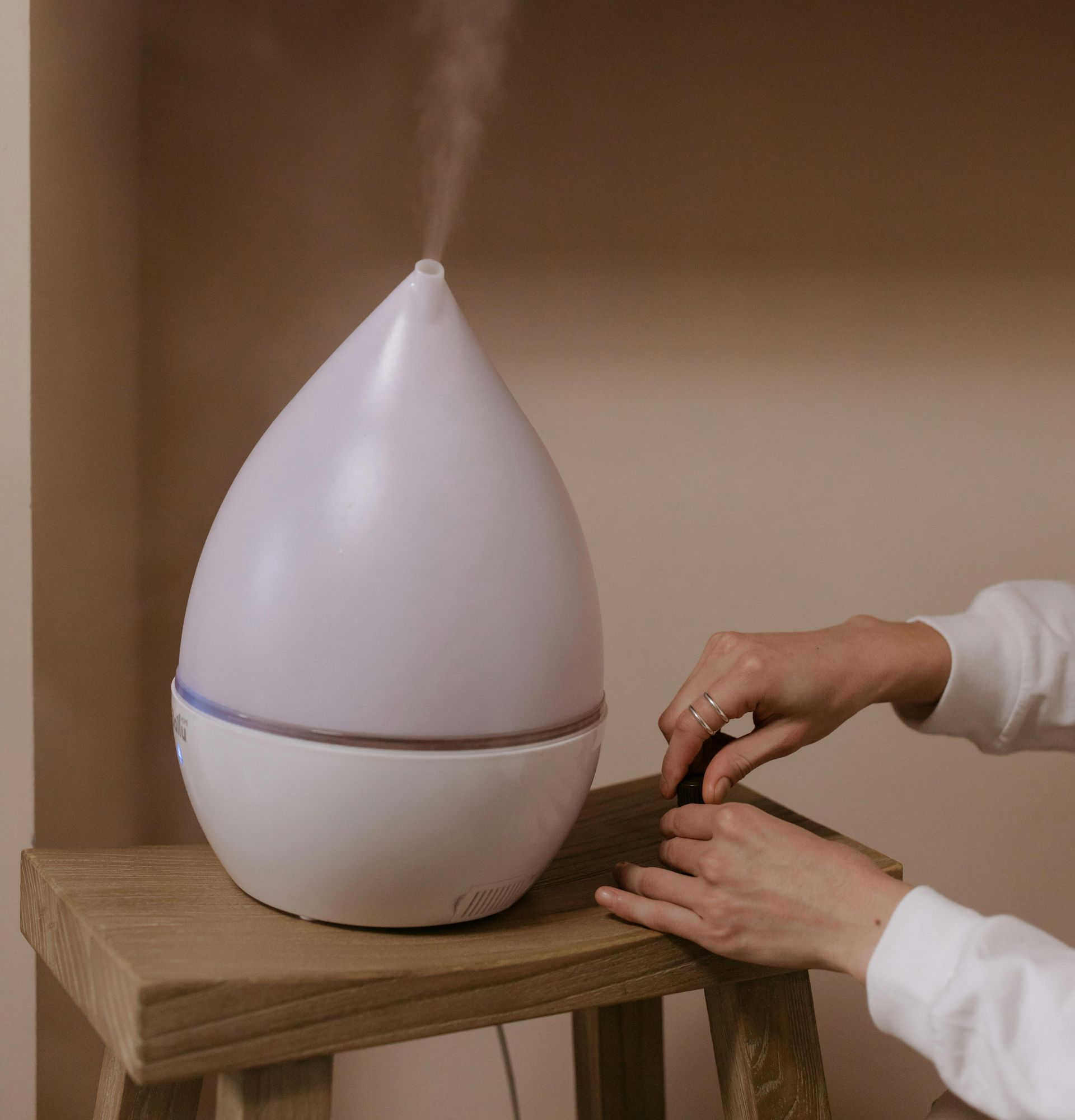 White teardrop-shaped diffuser emitting mist on a wooden table, with hands adjusting the base