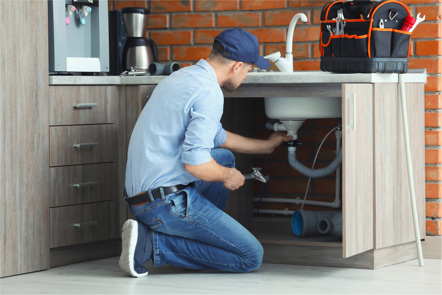 Plumber kneeling under a sink, inspecting pipes in a kitchen with gray cabinets and a brick backsplash