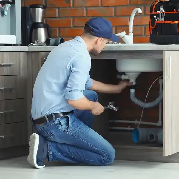 Plumber kneeling under a sink, inspecting pipes in a kitchen with gray cabinets and a brick backsplash