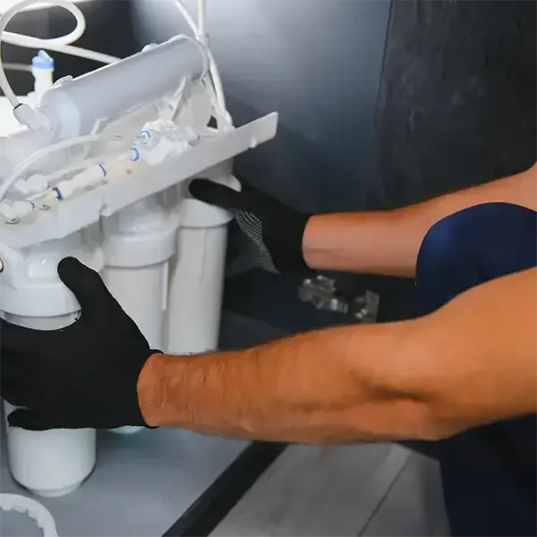 Person installing a white water filtration unit under a sink, wearing black gloves.