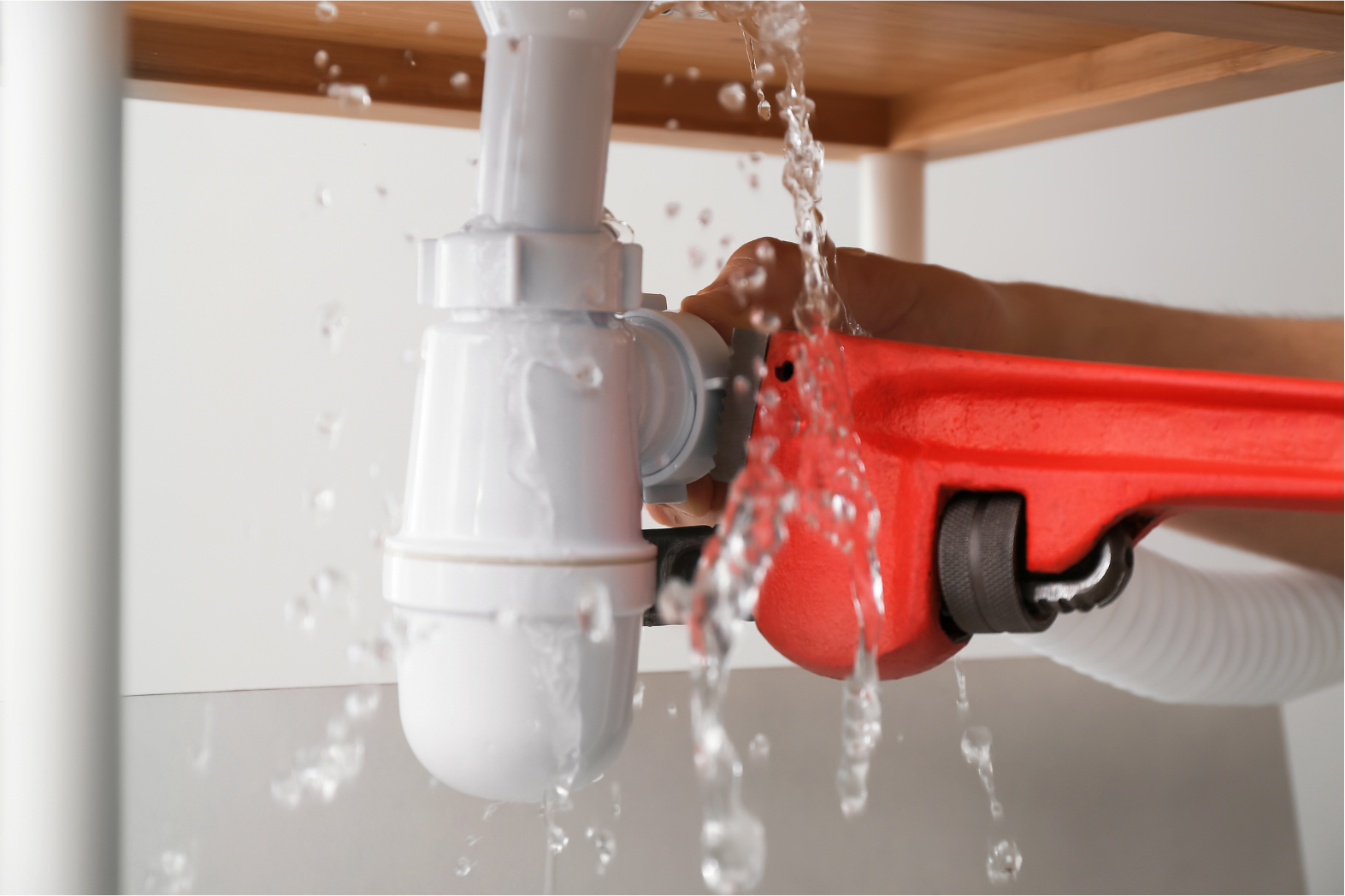 Water leaking from a sink drain pipe under a counter, with a hand holding a red pipe wrench nearby