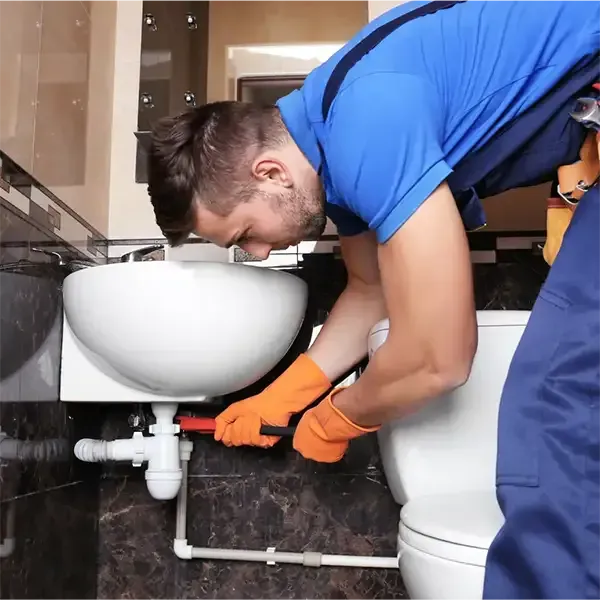 Plumber in blue shirt fixing pipes under a sink with orange gloves in a bathroom