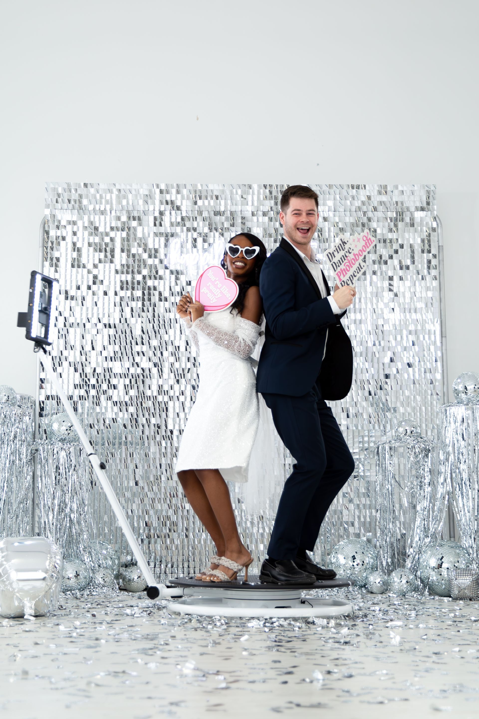 Two people in formal attire pose on a 360-degree photo booth platform in front of a shimmering silver sequin backdrop.