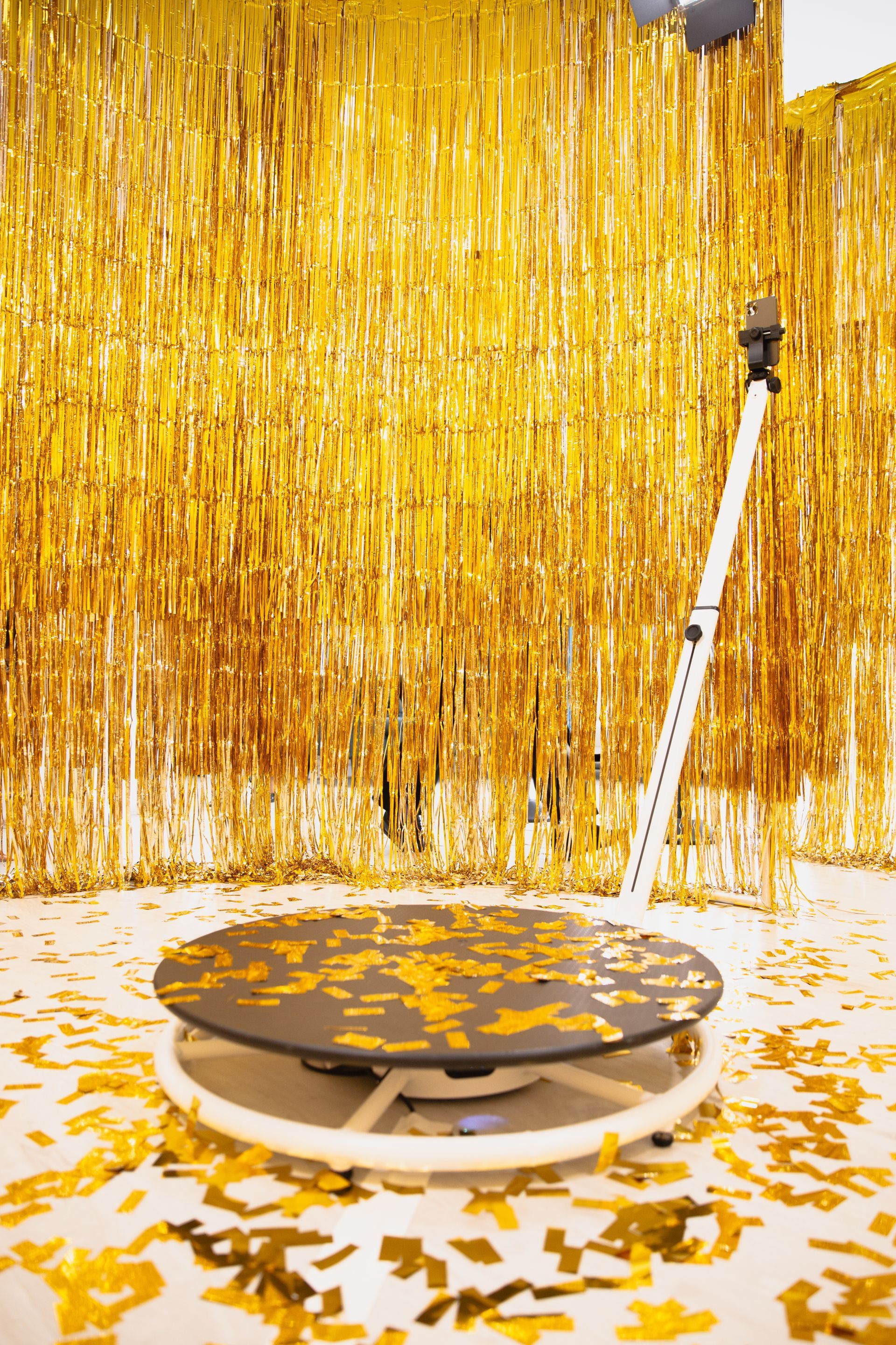 A 360-degree photo booth platform stands in front of a gold tinsel curtain, surrounded by scattered gold confetti.