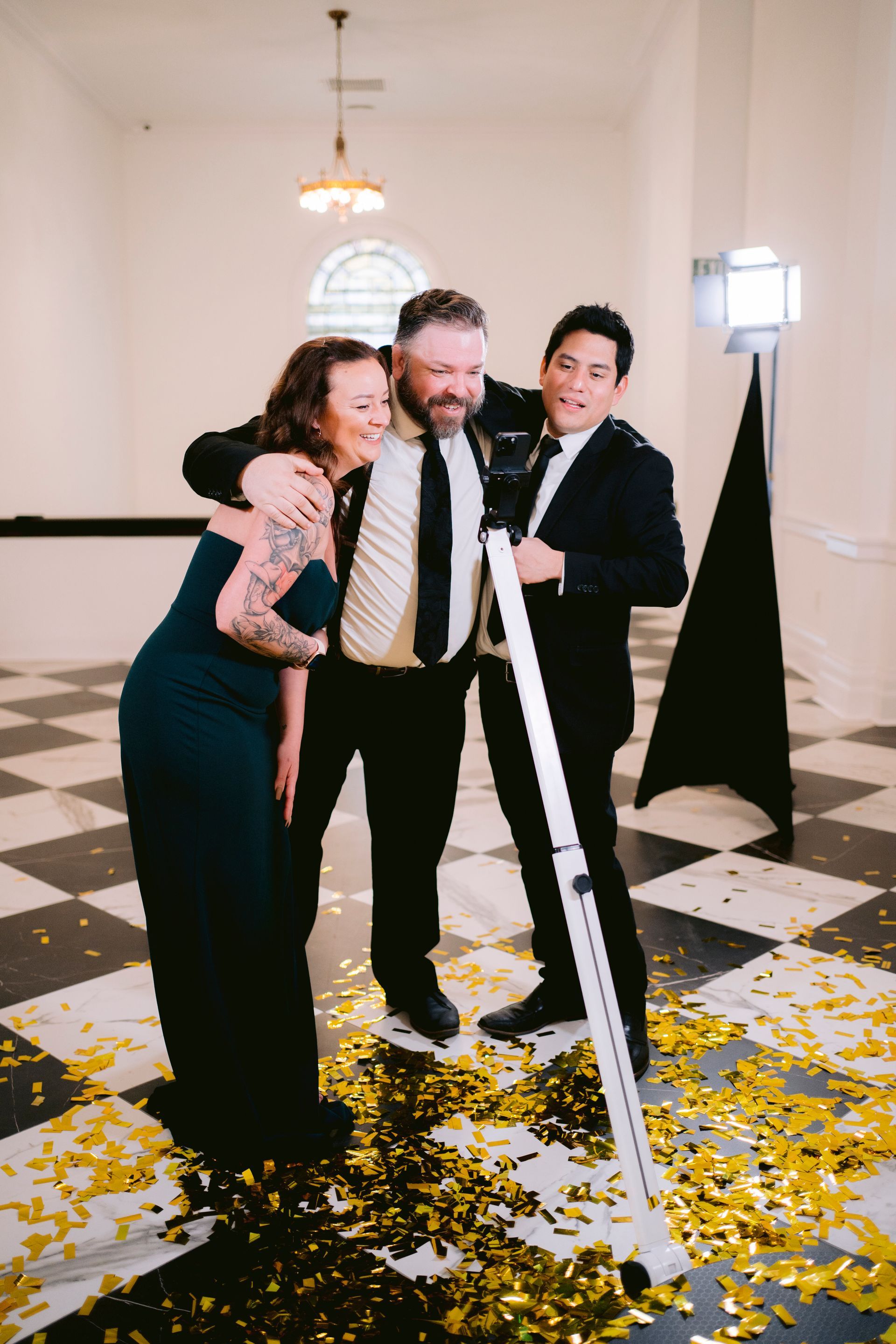 Three people stand together laughing on a checkered floor covered in gold confetti, near a light stand.