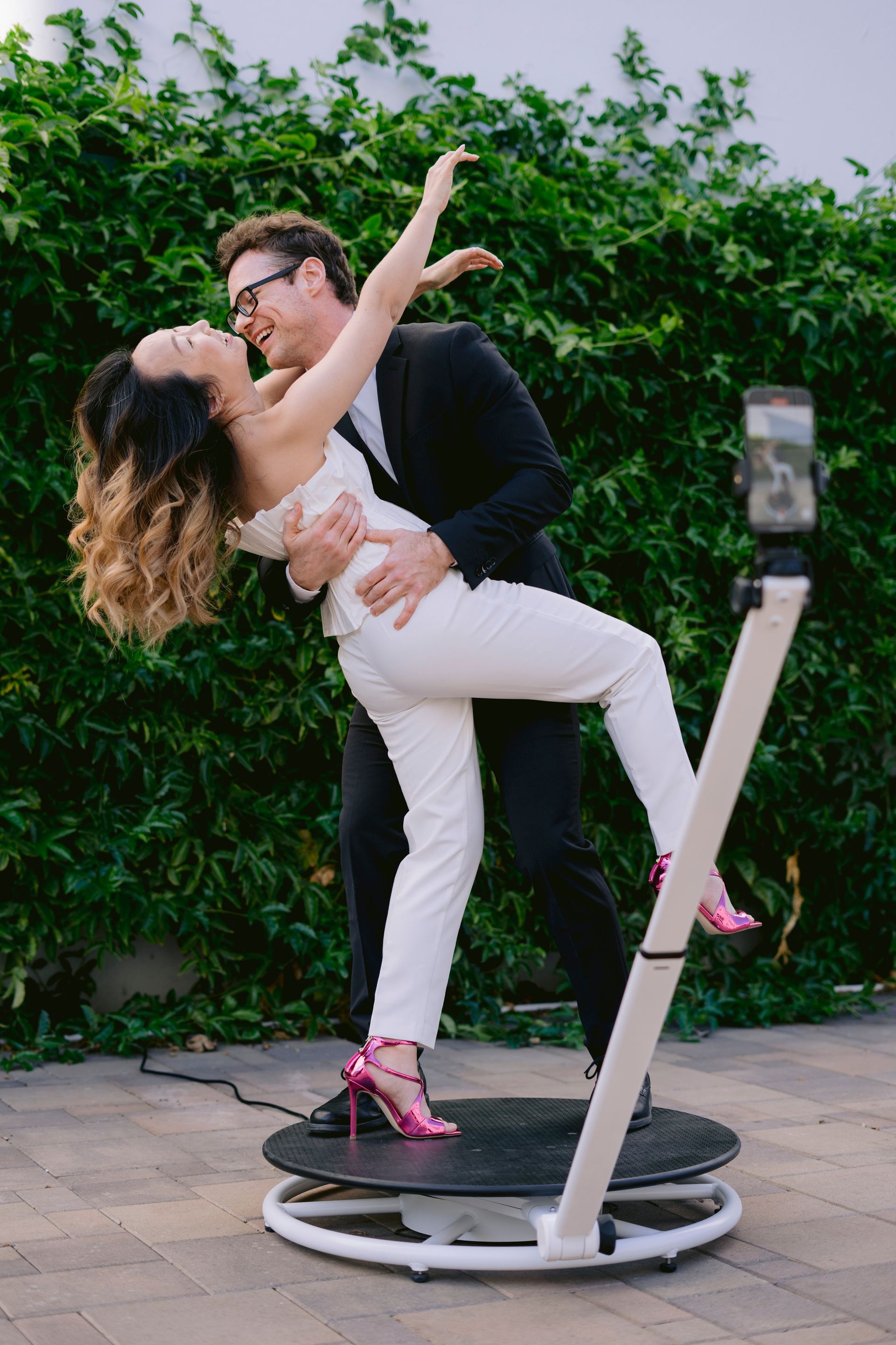 A couple in formal attire posing on a 360-degree rotating photo booth platform in front of a green hedge.