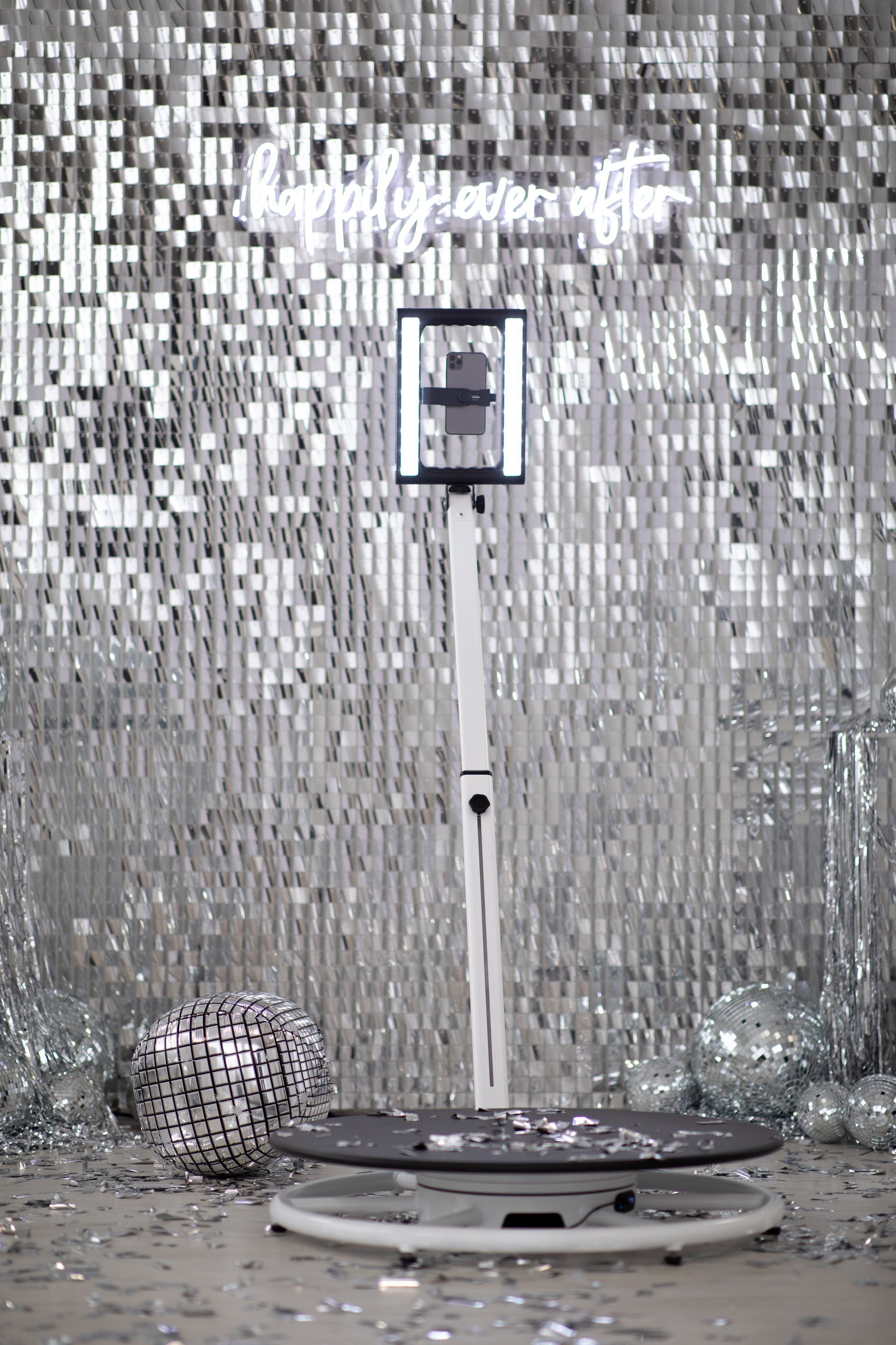 A 360-degree photo booth platform with a tall camera mount stands before a sparkling silver sequin backdrop.
