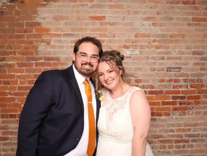 A couple stands smiling in front of a textured red brick wall. The man wears a navy suit with an orange tie.