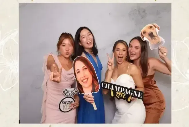 Four people in a photo booth pose with fun props including a dog face cutout, a diamond ring sign, and a champagne sign.