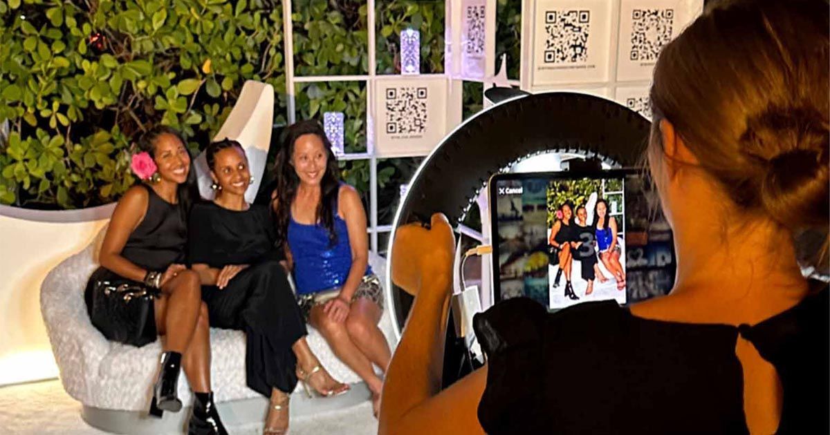 Three women posing on a couch for a photo booth session, viewed from behind the photographer holding the ring light.