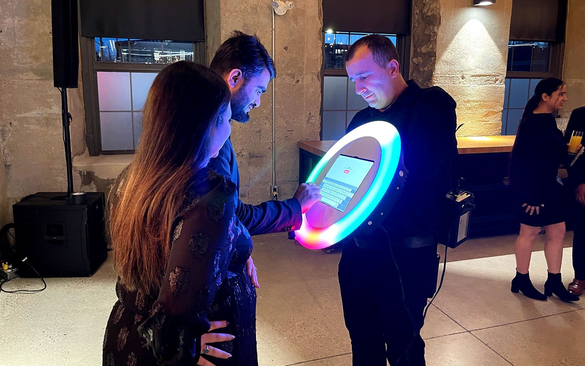 People interact with a glowing circular ring light photo booth in an indoor event space with stone walls.