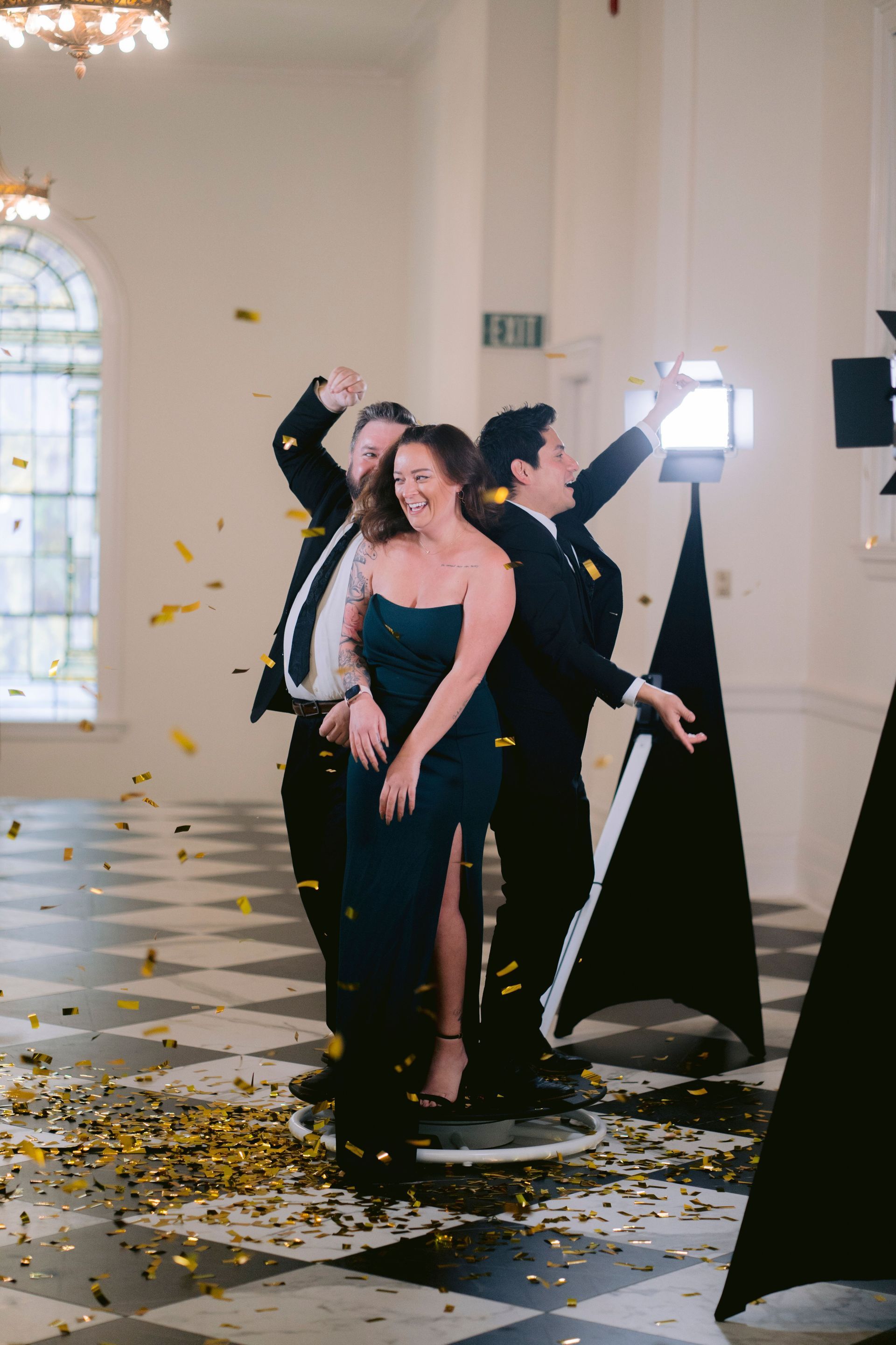 Three people pose on a spinning platform during a 360-degree video booth session while gold confetti falls around them.