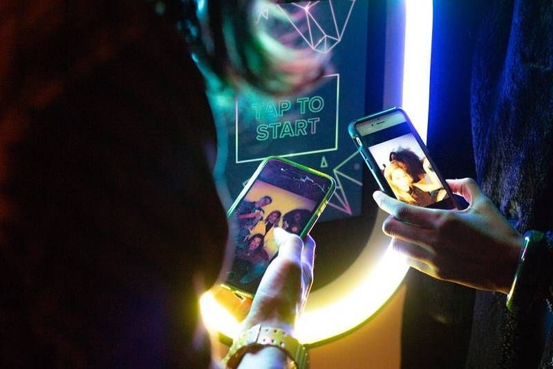 Two people holding smartphones showing photos in front of a brightly lit, circular photo booth display.