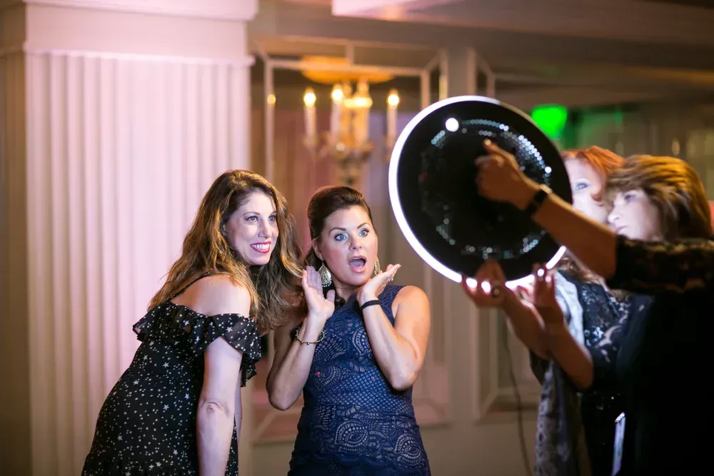 Two people pose for a photo with a light ring held by a person in the foreground at a party.