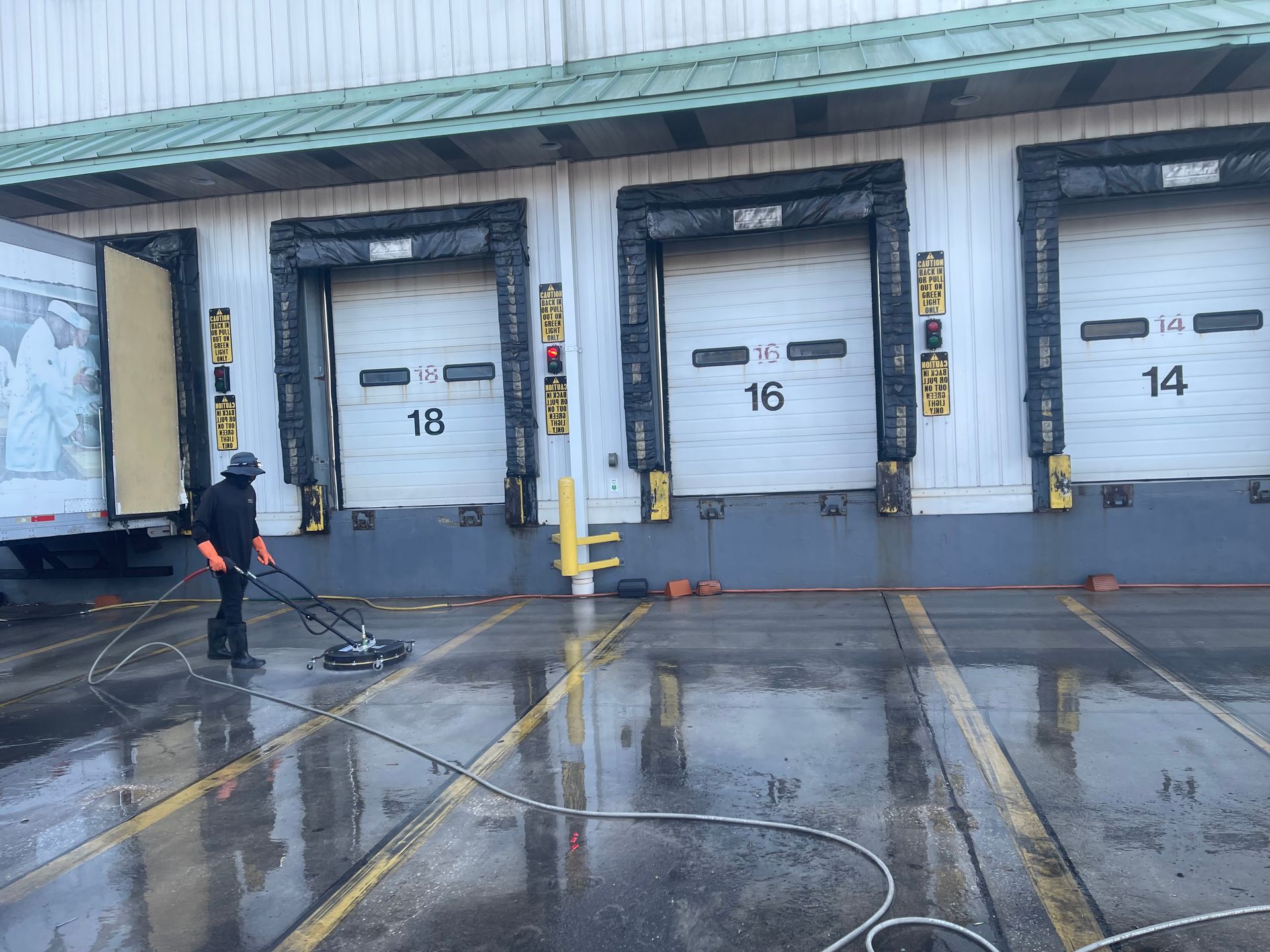 Worker pressure-washing loading docks outside a warehouse with numbered bay doors and wet pavement