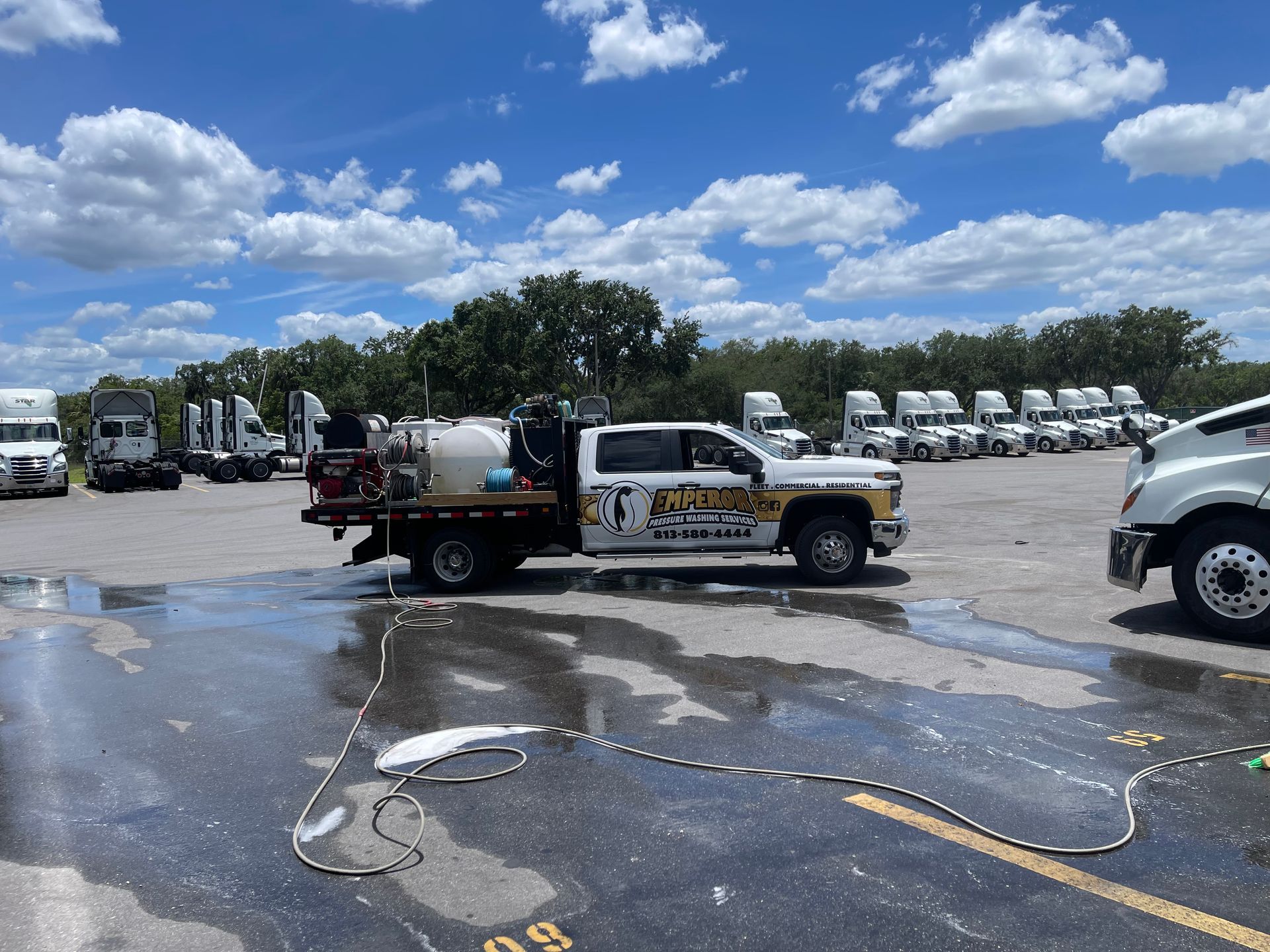 Work truck with tank in a parking lot beside rows of white trucks under a blue sky.