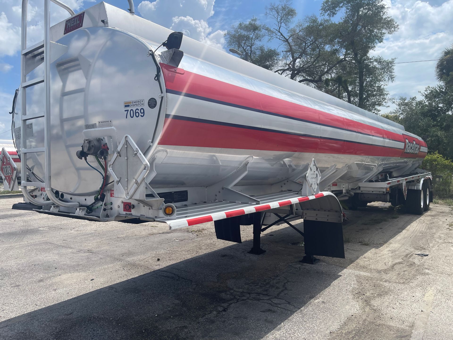 Silver tanker trailer with red stripe parked outdoors on a gravel lot under a cloudy sky