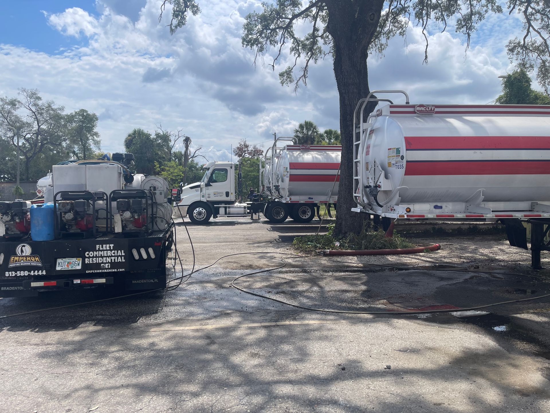 White tanker trucks and a tow truck parked on a muddy lot under a large tree.