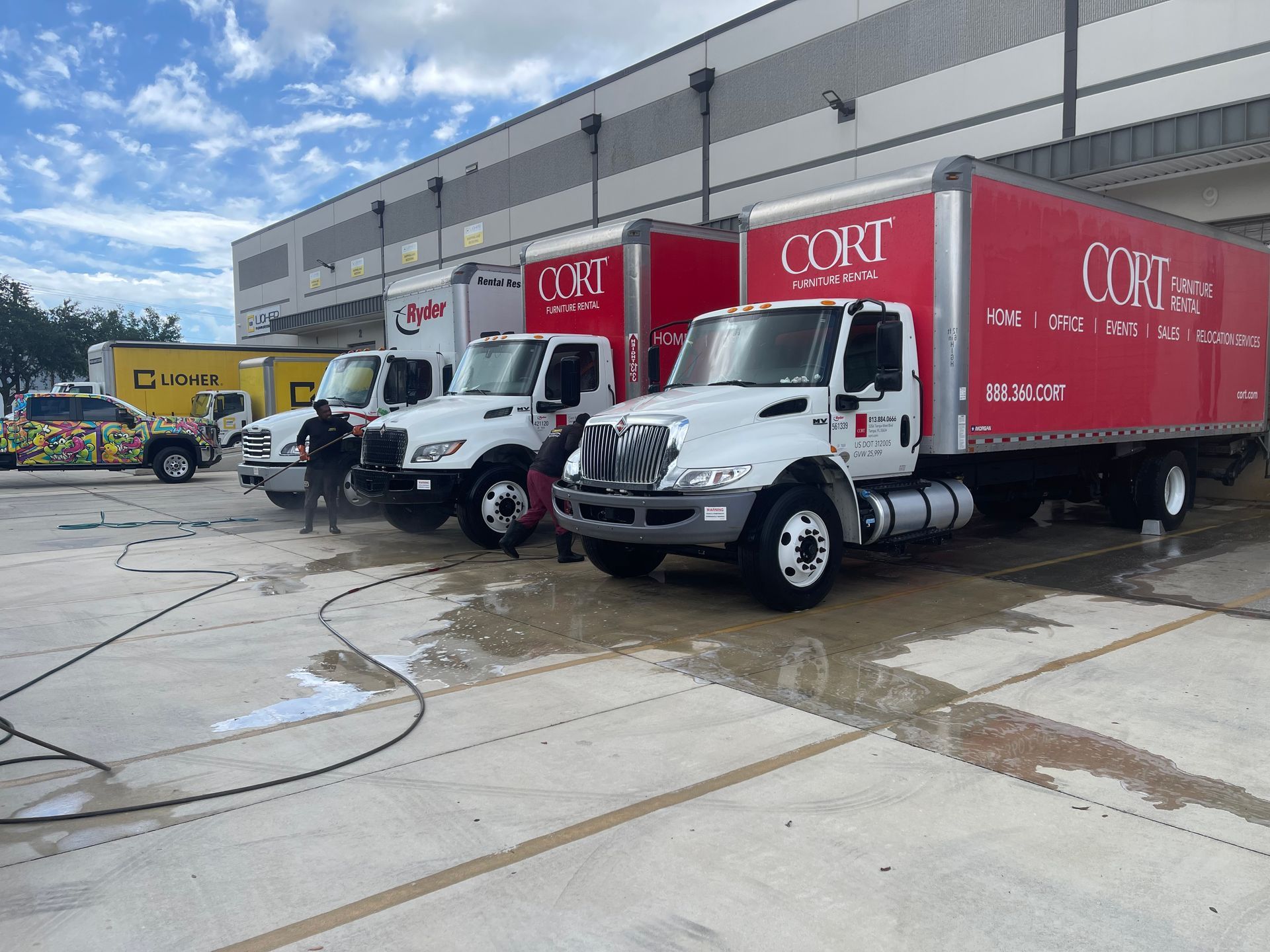 Delivery trucks lined up at a warehouse dock on a wet concrete lot.