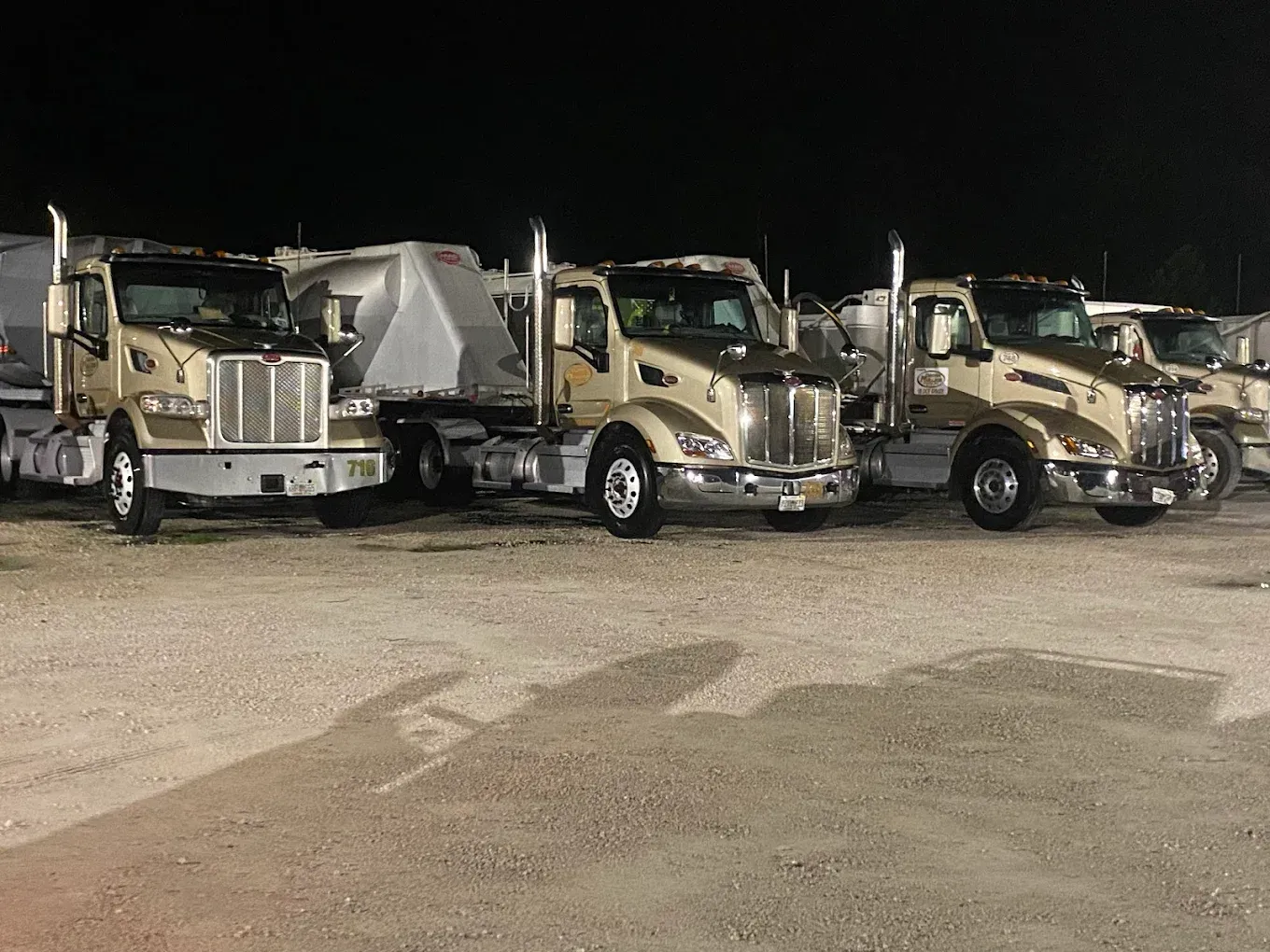 Row of beige semi-trucks parked side by side in a lot at night