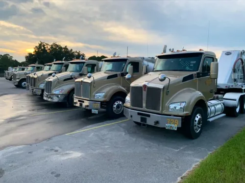 Row of beige semi trucks parked along a road at dawn, with a cloudy sunrise in the background