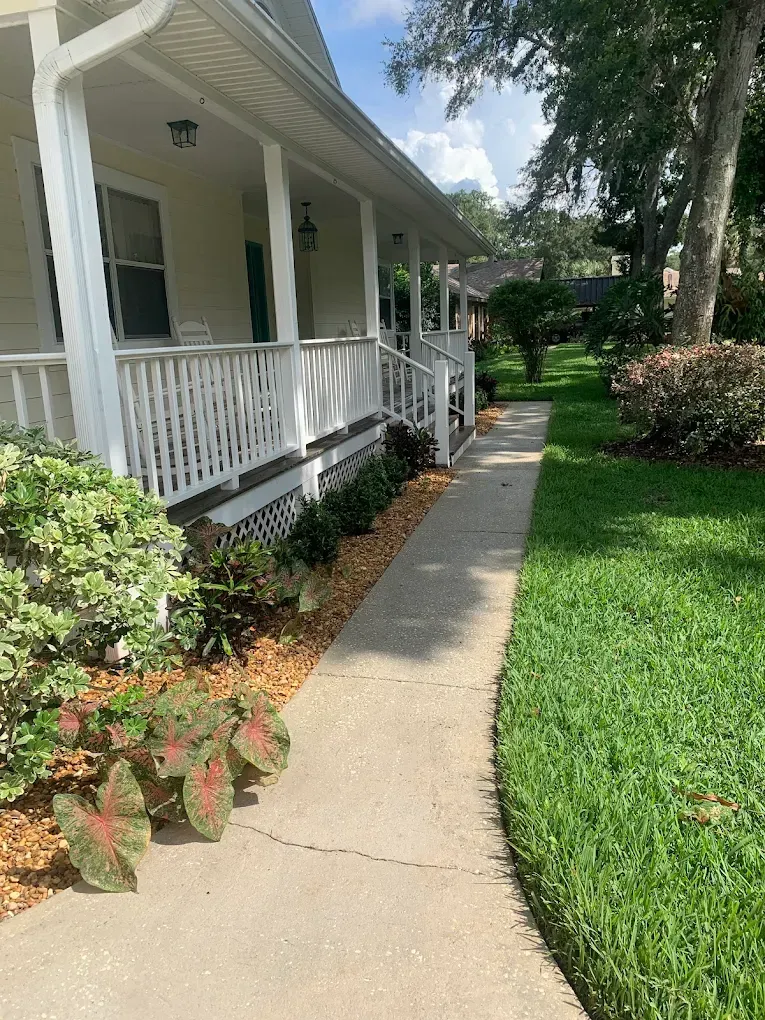 Front porch with white railing and potted plants beside a concrete walkway in a green yard