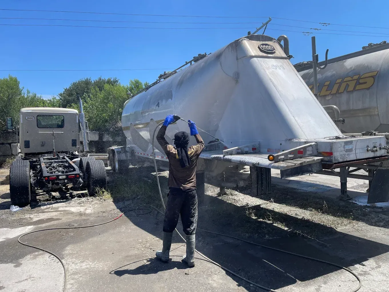 Person washing a large white boat trailer in a sunny lot, with trucks and a boat nearby