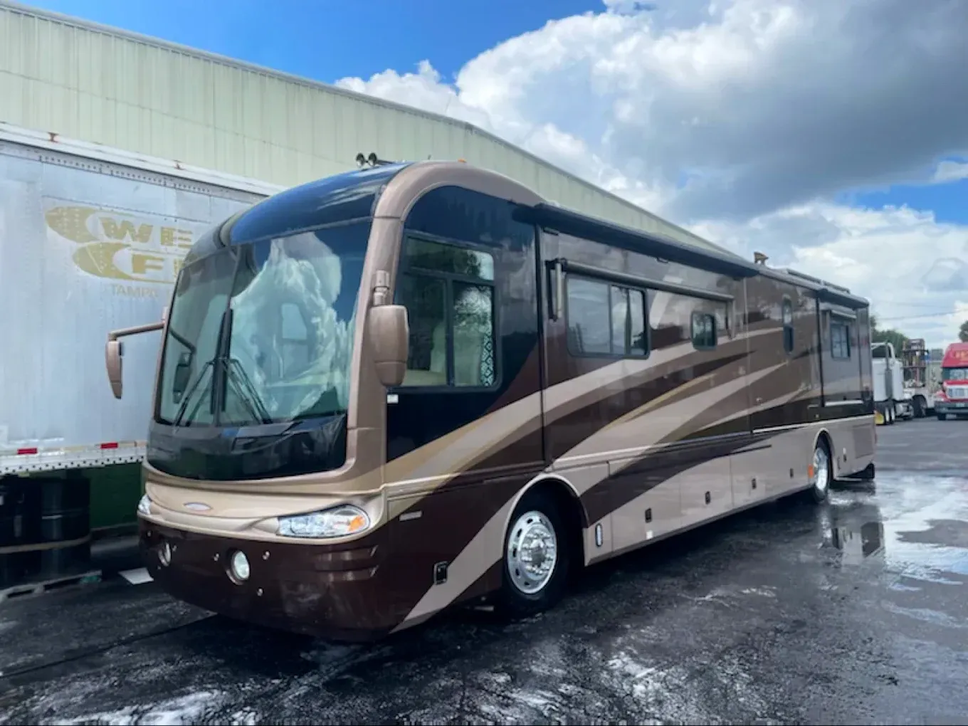 Brown and beige luxury coach bus parked on a wet lot under a cloudy sky