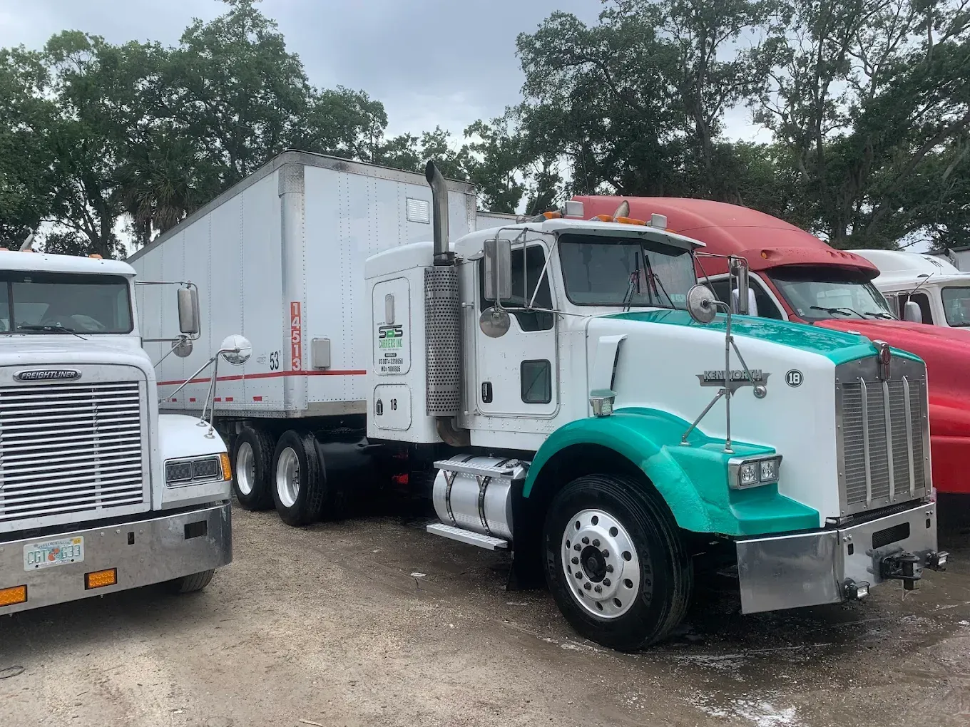 White and teal semi-truck parked in a gravel lot beside another truck, with trees in the background