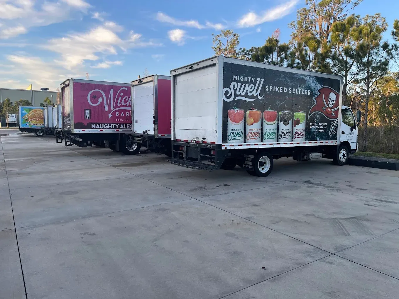 Food trucks parked on a lot at sunset, including a pink truck and a truck with colorful drink graphics.
