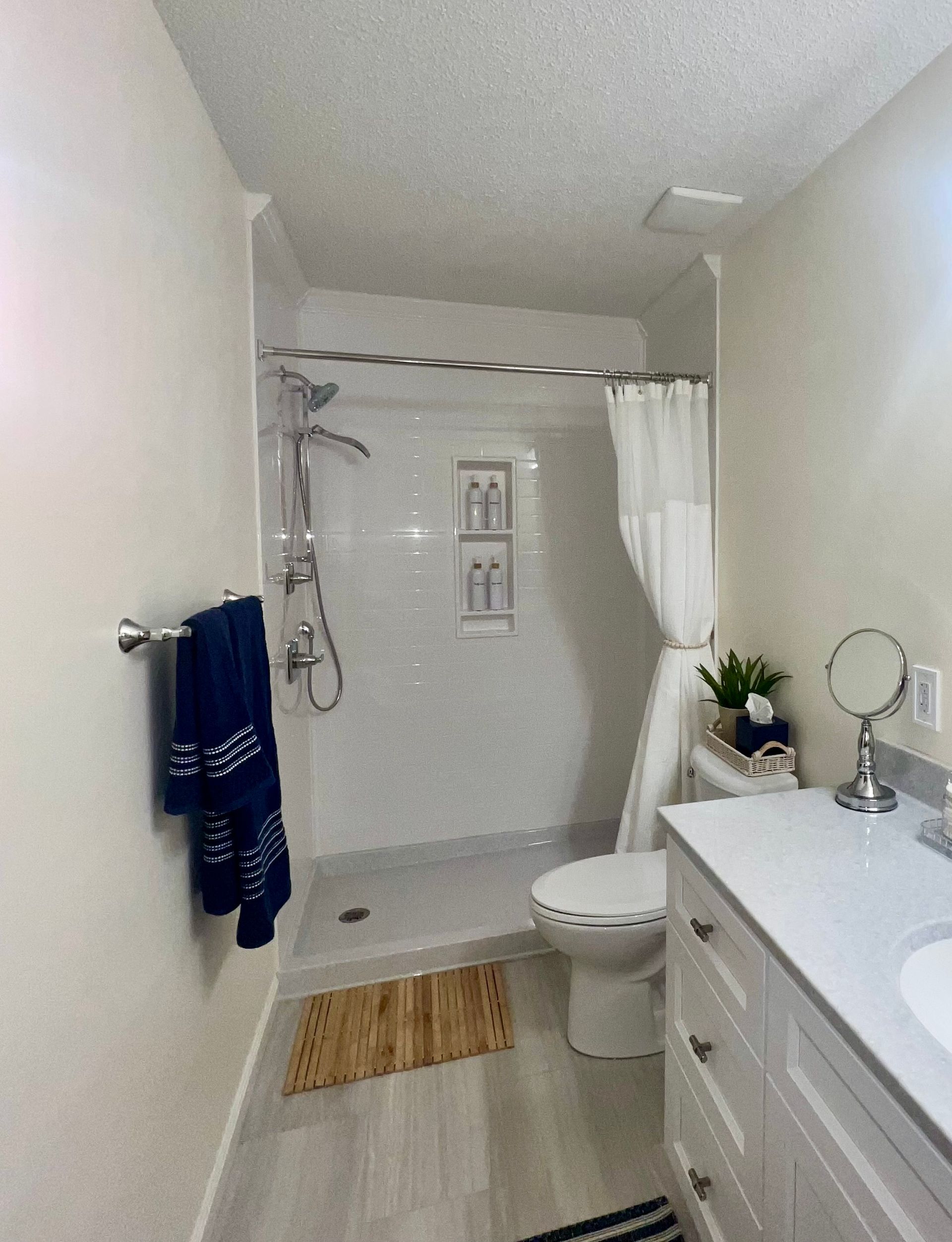 Narrow white bathroom with walk-in shower, toilet, sink vanity, mirror, towel, and small rug.