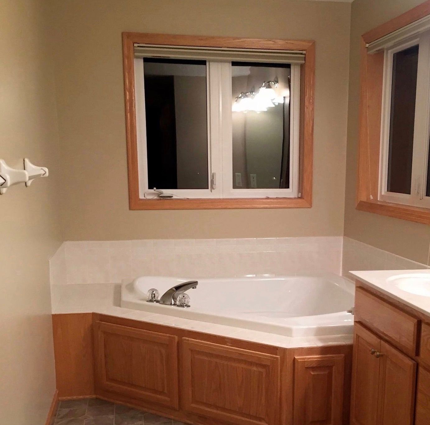 Empty bathroom with a corner tub, wood cabinets, and two windows at night.