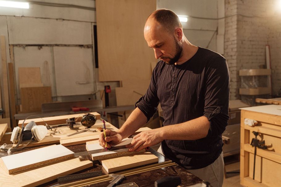 Man working with wood in a workshop, measuring boards on a cluttered workbench.