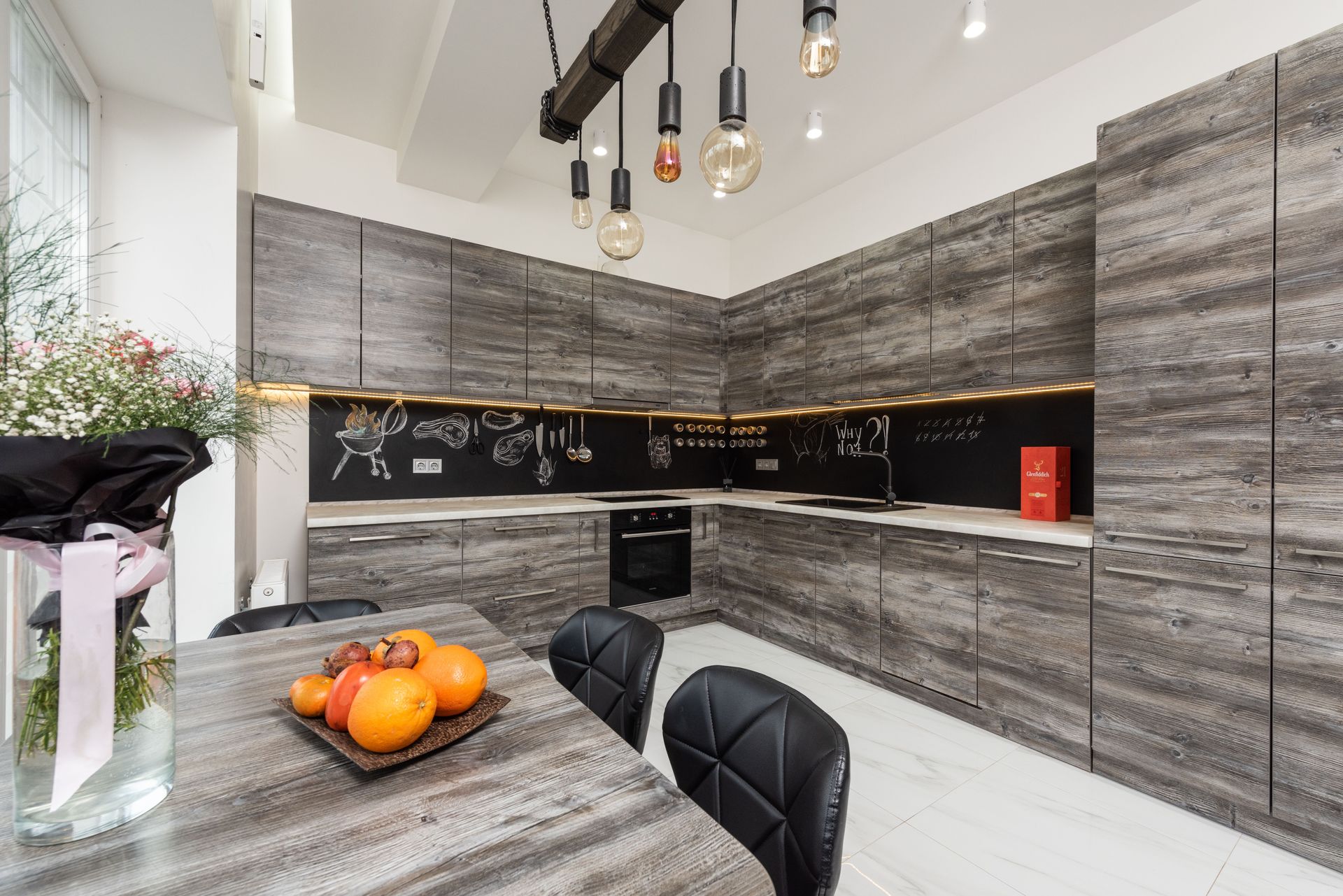 Modern gray kitchen with black backsplash, marble island, hanging lights, and bowl of oranges