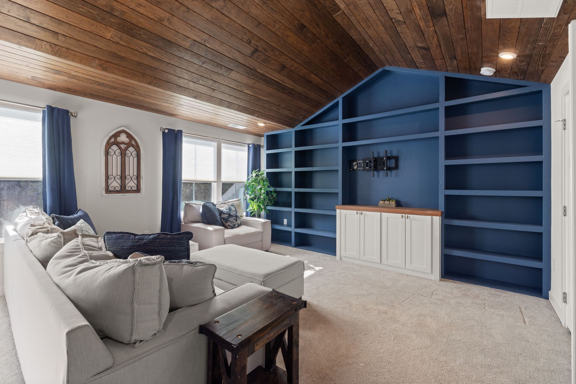 Spacious living room with a vaulted wood ceiling, beige sectional, and large built-in blue shelving wall