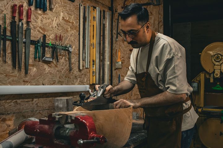 Woodworker shaping wood at a bench in a workshop, surrounded by hanging hand tools.
