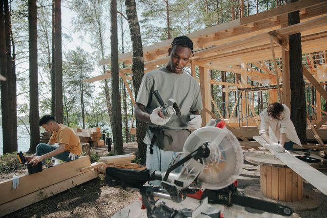 People building a wooden structure in a forest workshop, with a saw in the foreground.