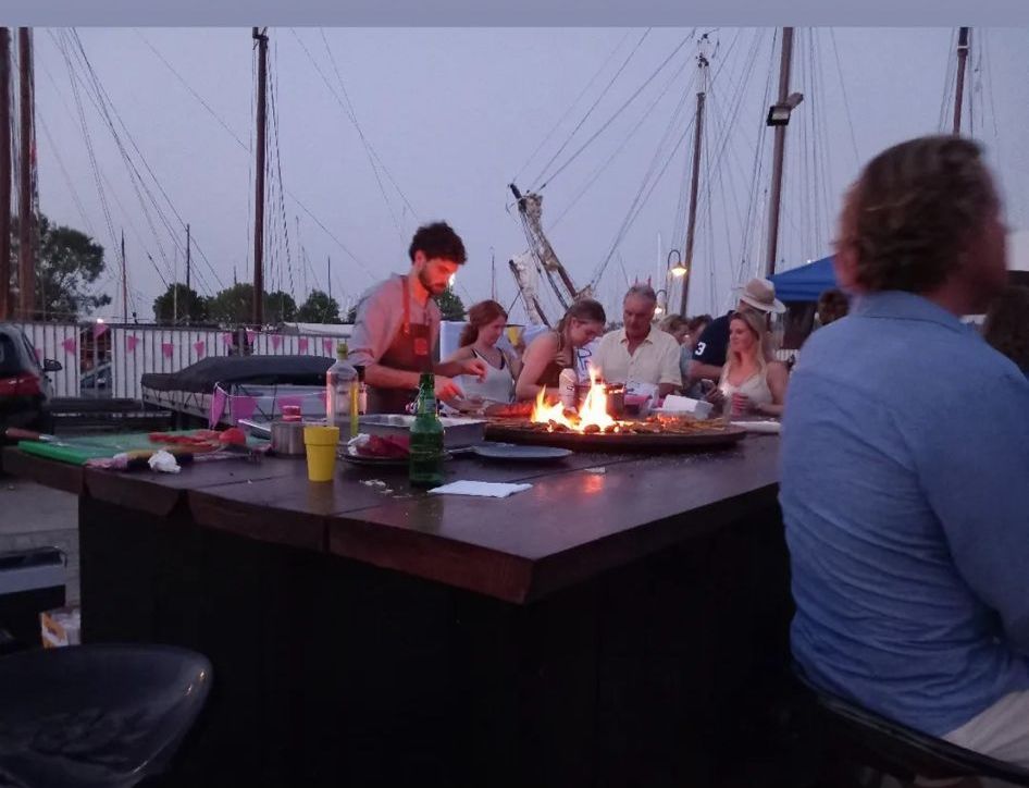 Mensen zitten rond een tafel met eten, dat wordt bediend door een man met een vuurkorf, met masten op de achtergrond.